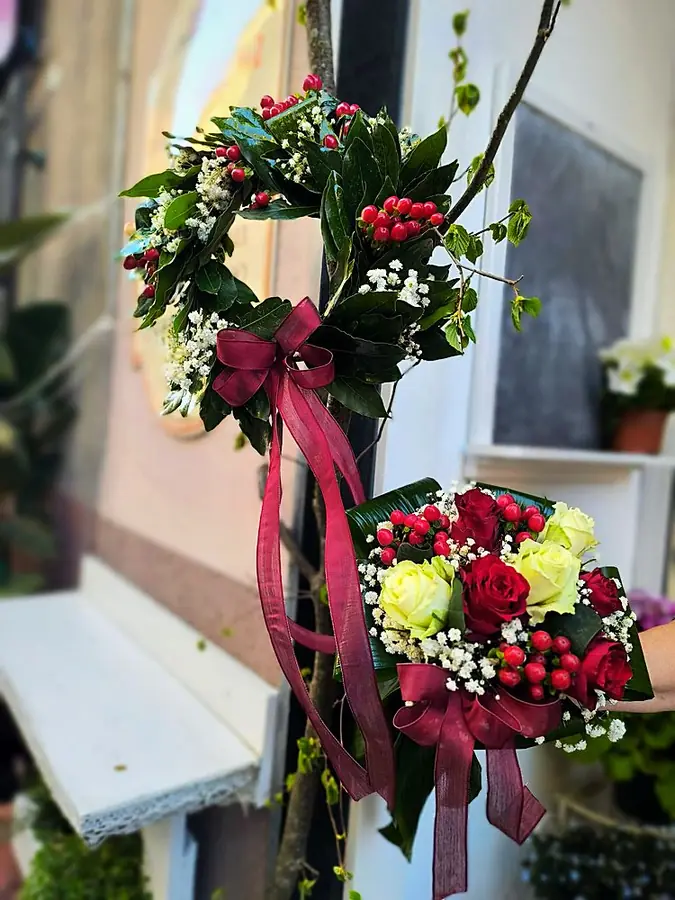 Corona d’alloro con bacche rosse e nastro bordeaux, accanto a un bouquet di rose rosse e bianche