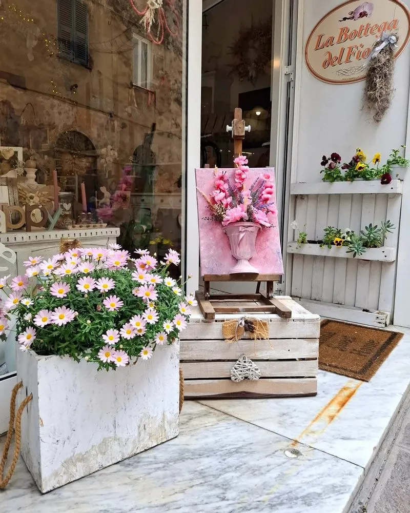 Ingresso de La Bottega del Fiore con margherite rosa, quadro floreale su cavalletto e vasetti di viole