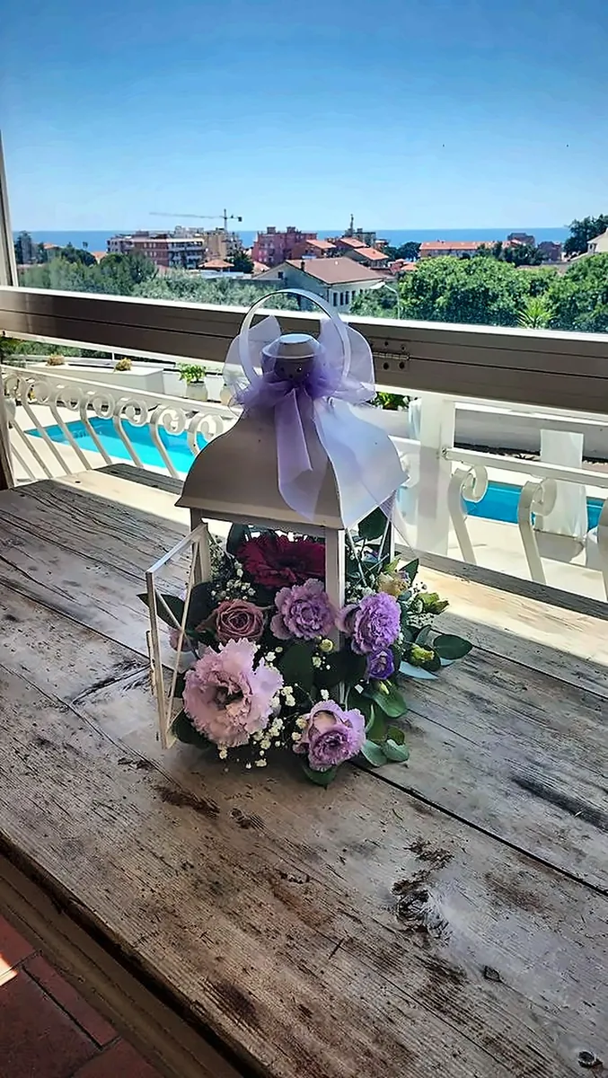 Lanterna bianca con fiori lilla e fiocco in organza, centrotavola su tavolo in legno con vista sul mare