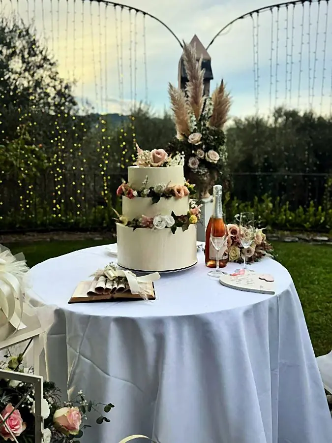 Torta nuziale a tre piani con fiori freschi e pampas, accanto a spumante e bicchieri su un tavolo in giardino