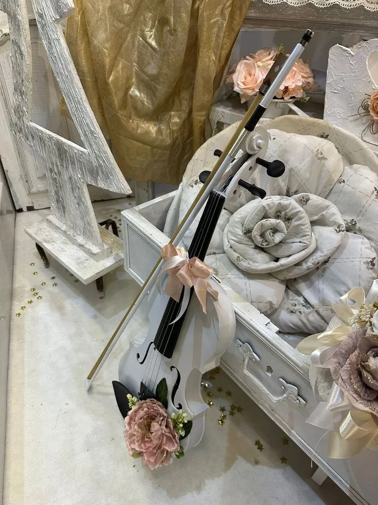 Violino bianco decorato con peonia rosa e nastro color pesca, appoggiato a un cassetto d’epoca con cuscini a fiori